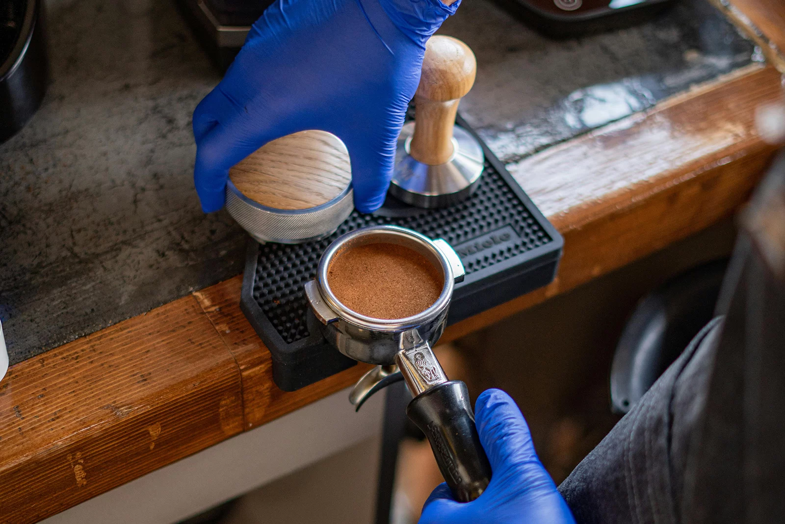 Barista w niebieskich gumowych rękawiczkach ubija kawę do kolby na kawę.