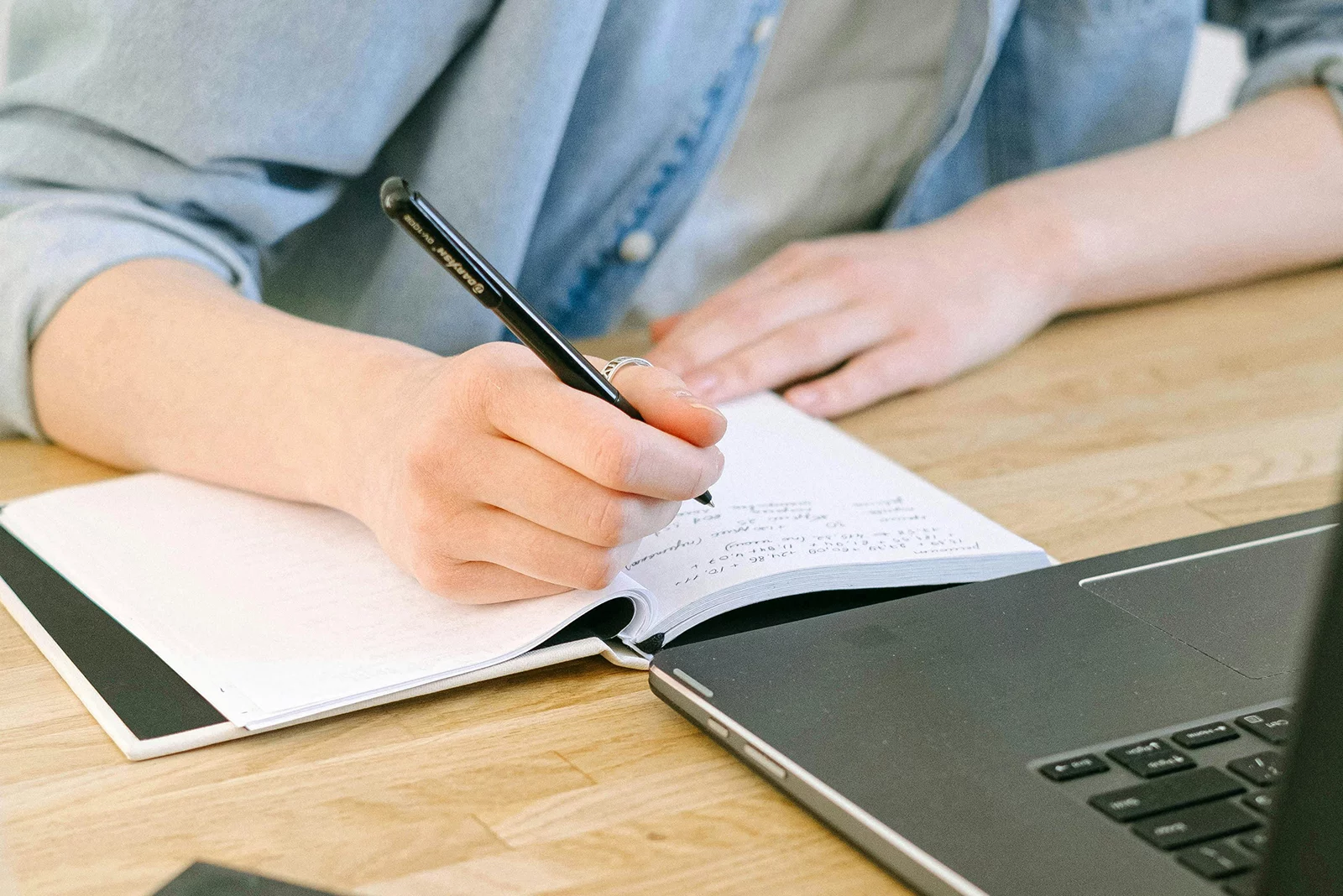 Osoba siedząca przy biurku, notuje coś w notatniku z ekranu laptopa.